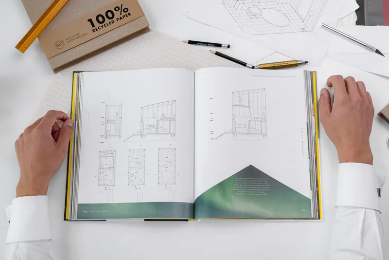 The book open on a desk of drawings and pencils, two hands on its pages