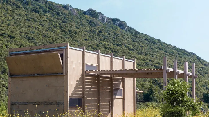 The plywood cabin in a meadow below a green hillside, a shutter propped open at one end and a slatted pergola at the other