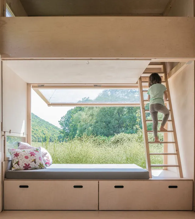 Inside: a cushioned bench under the big window, swung up like a canopy over the meadow, and a child climbing the ladder to the loft