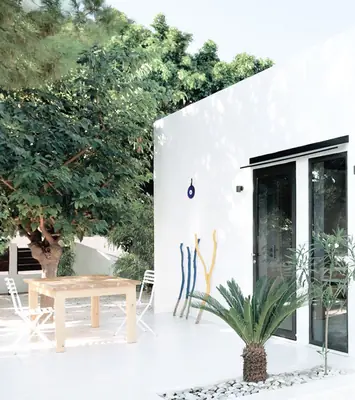 The white Monocabin with black glass doors, a table and a palm on its white terrace under a tree