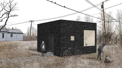 A black shingled cube on an empty lot beside a white house, power lines overhead