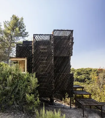 A dark timber cabin wrapped in slatted panels on a wooded slope, steps leading up to it