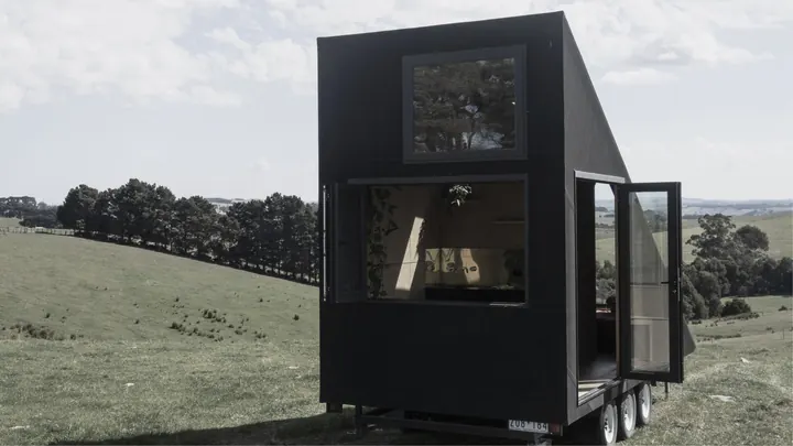 The black Base Cabin on its trailer on a green hillside, a high window in its end wall and its glass door open