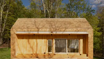 A timber-clad micro home with a wide glass front at the edge of a forest