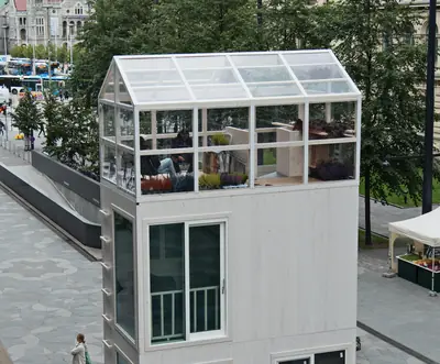 A slim white timber tower with a glass greenhouse on top, on a city square