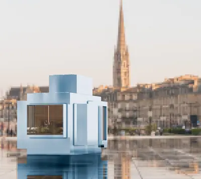 A pale blue cube house on the water, a church spire and old city behind