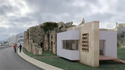 A white box set against the stone walls of a ruin, by a road along the sea