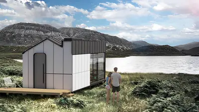 A white panelled cabin with a gabled roof by a mountain lake, two people looking at it