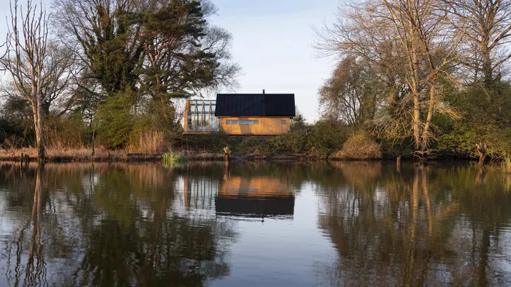 Cabin ANNA seen across a still lake at dusk, a lit window between the trees and its reflection in the water