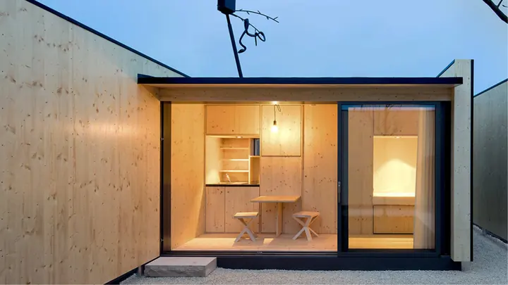 The Micro Courtyardhouse at dusk: pale spruce walls around a small gravel courtyard, and through the long glass door a lit room with a table and two stools