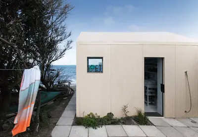 The pale metal-clad house by a paved path, a towel on the line and the sea behind