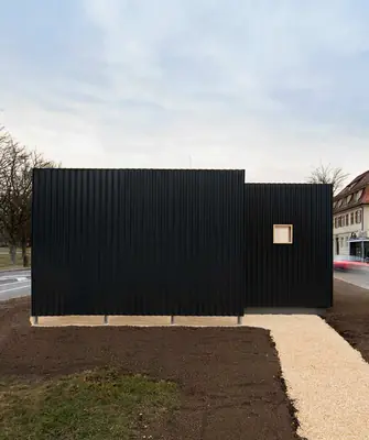 A low black box of corrugated metal on a gravel island beside a road, houses behind