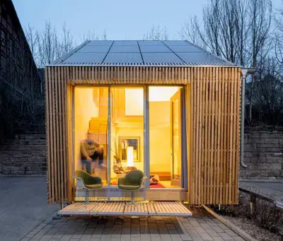 A small timber-slatted house lit at dusk on a paved car park, people inside
