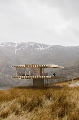 A timber house on stilts with a deep flat roof on a golden grassy hillside, mountains behind