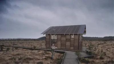 A shingled cabin at the end of a boardwalk across a dark heath under a grey sky