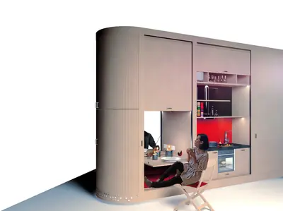 A rounded pale unit with a kitchen, shelves and a table built in, a woman seated at it