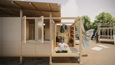A woman sits cross-legged on the raised timber floor of an open shelter, a curtain blowing beside her