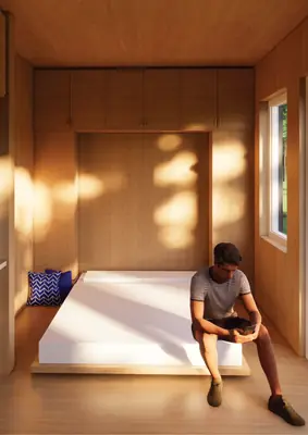 A man sitting on a bed in a timber room lit by low sun through the window