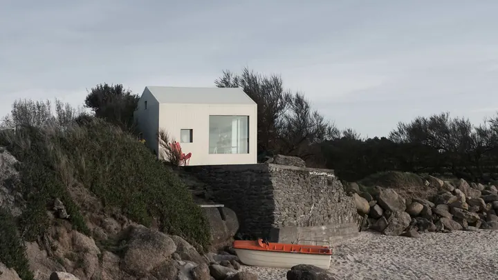 The small white house on a stone base above a rocky beach, shrubs around it and a boat on the sand
