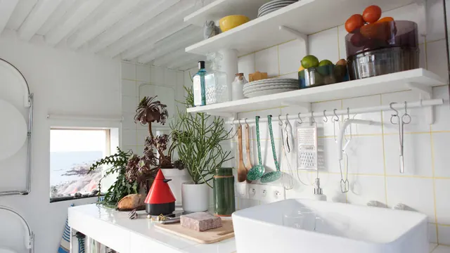 The kitchen: white shelves of plates, fruit and plants over a white sink, tiles with yellow joints behind