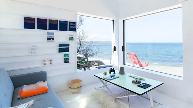 Inside: a grey sofa and a low table by a corner of big windows onto the beach and the sea