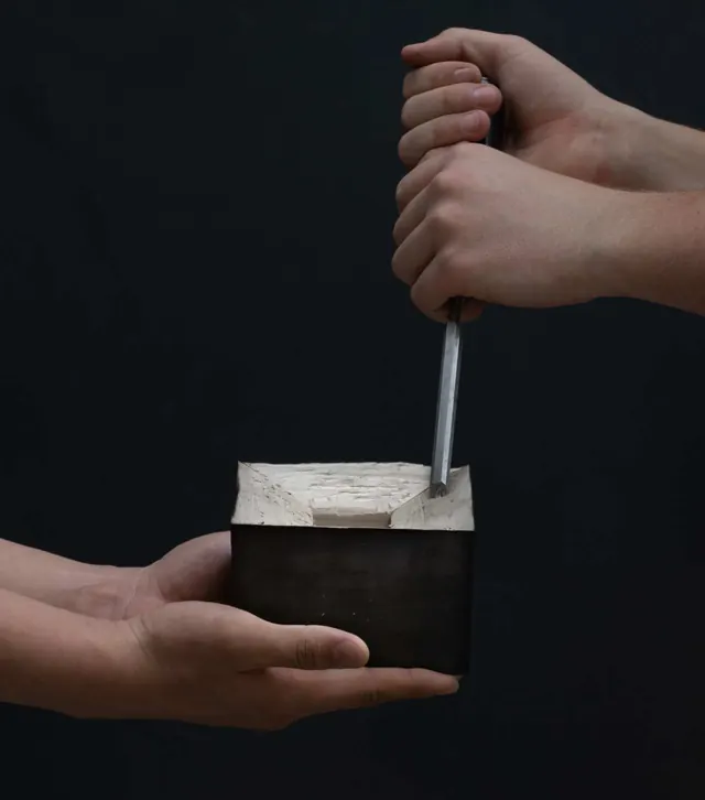 Two hands carving a small block model of the house with a steel tool, on black