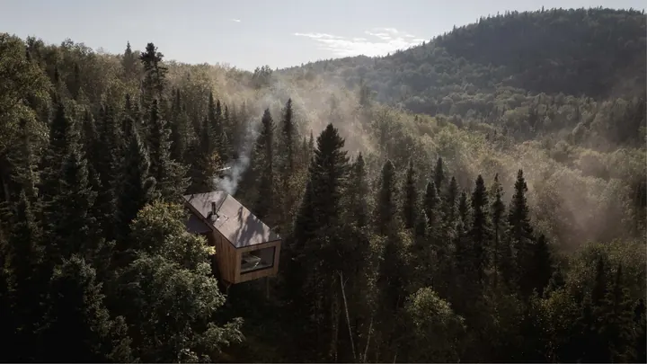 A small timber shelter on legs in a steep forest of dark firs, smoke rising from its chimney into the mist