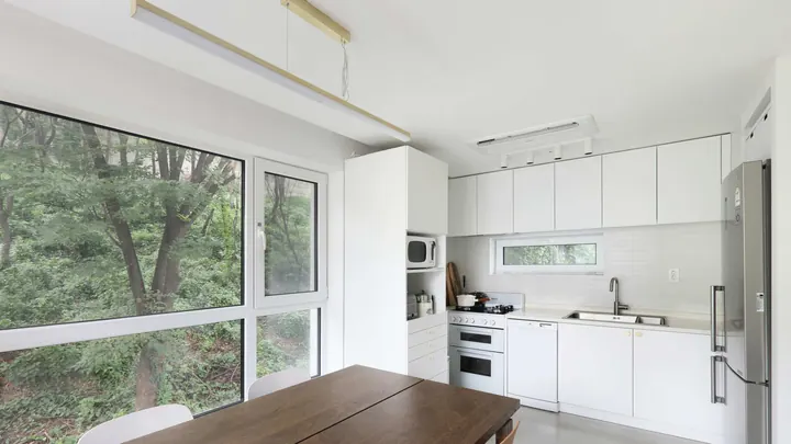 The kitchen floor: white cupboards, an oven and a fridge, a wooden table, and wide windows onto the trees