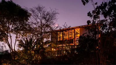 The house at dusk among trees, its long timber front lit from inside under a violet sky