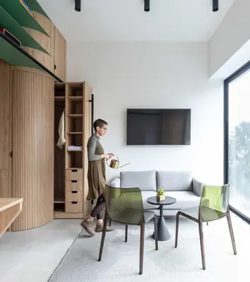 A white studio with a tall timber wall of cupboards, a man by a small table with green chairs