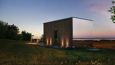 A box house of timber and mirror glass on a grassy shore at dusk, lights at its foot