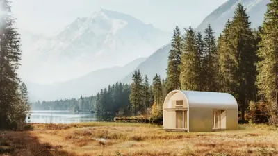A curved timber cabin in a meadow by a mountain lake, pines behind it