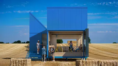 A blue house open to a wheat field, people on its steps and in the open room