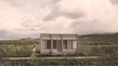 A long timber box house on a grassy plain under a cloudy sky, a woman walking past