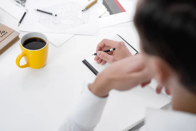 Someone writing in the notebook, head in hand, a yellow mug of coffee close by