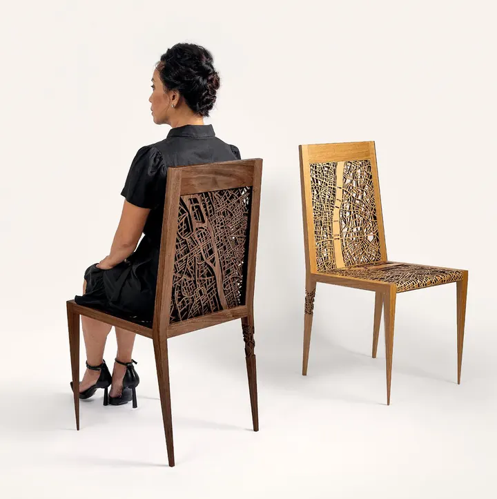 A woman seated on the Placemaking Chair: Paris, beside a second chair, both with laser-cut map backs