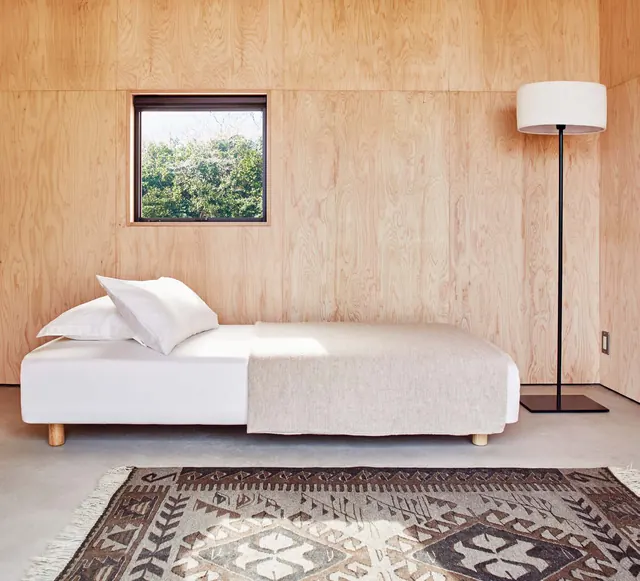Plywood walls, a white bed, a floor lamp and a small square window