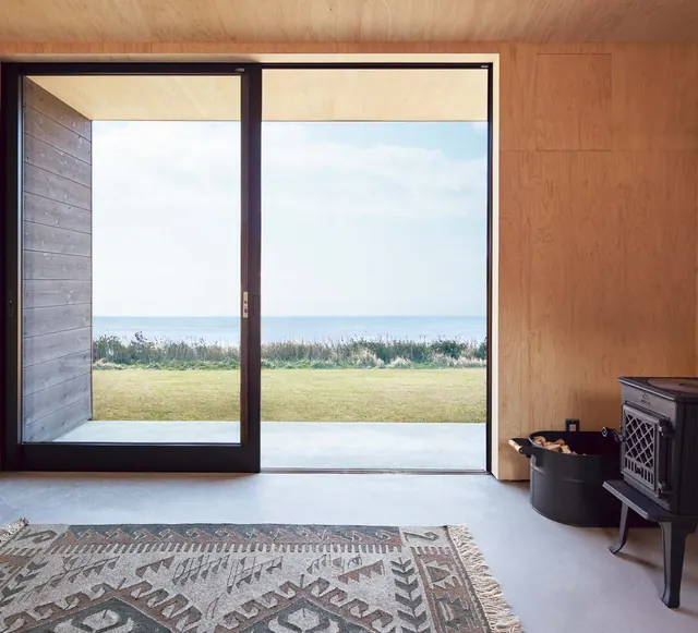 Inside the hut: the sliding glass door onto the grass and the sea, a wood stove and a rug