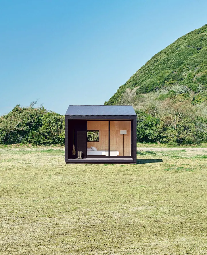 The small black hut on a meadow, its glass door open, a green hill behind