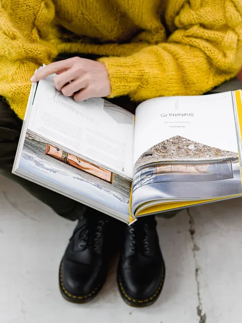 El libro abierto sobre las rodillas de quien lee, arriba un suéter amarillo y abajo unas botas negras