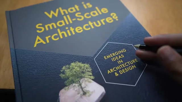 The cover of the book on a desk, a hand pointing a pen at its hexagon