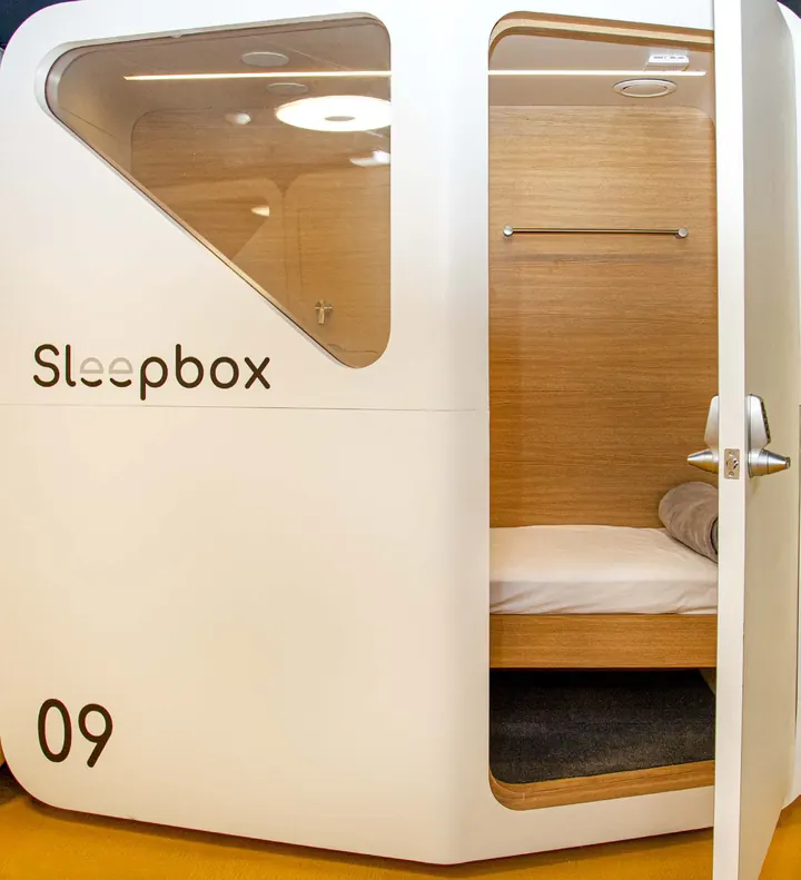 Una habitación blanca de Sleepbox con la puerta abierta que deja ver una cama, y encima una ventana redondeada
