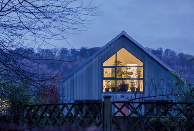 The house at dusk behind a garden fence, its gable window lit