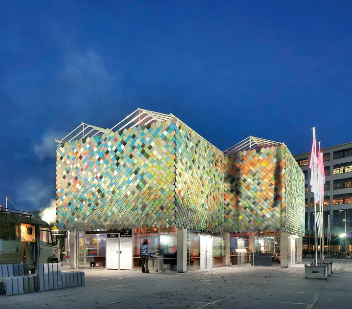 The pavilion at dusk, its upper façade a patchwork of coloured plastic tiles
