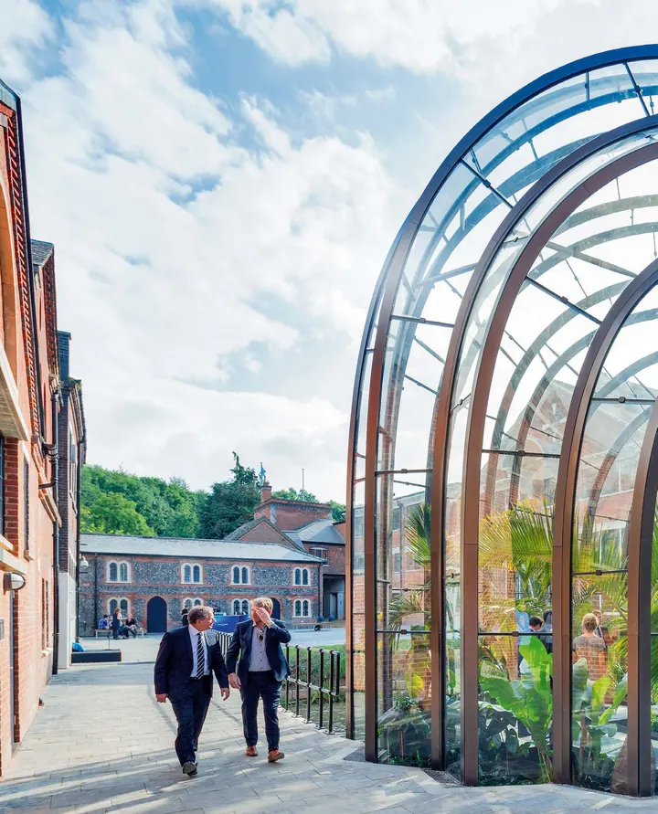 A curved glasshouse of bent steel ribs beside the old brick mill, two visitors walking past